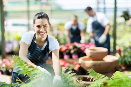 A young woman with dark hair pulled back, wearing a striped apron over a white t-shirt and light gardening gloves, is smiling while tending to green ferns in a bright greenhouse. Behind her, there are blurred figures of two people, likely gardening staff, wearing dark aprons and white shirts, working among a variety of flowering plants and potted greenery. The greenhouse has a glass roof allowing natural light to illuminate the lush plants, creating a vibrant and healthy garden environment. In the background, there are stacked stone bowls and neatly arranged flower beds, with the overall scene showcasing a well-maintained, professional gardening space that could be part of a local horticultural service provider in Hampstead, London, emphasizing their expertise in outdoor plant care and garden maintenance.